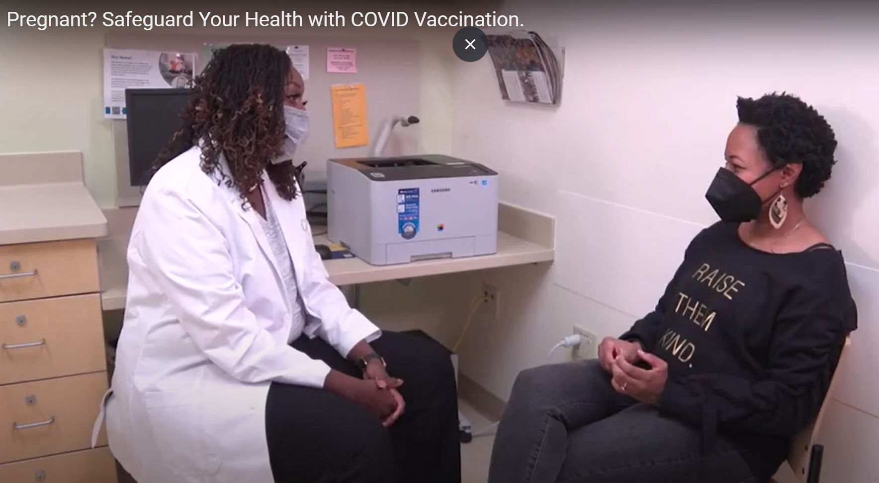 Two Black women wearing masks sit in a doctors office and talk. One is a doctor wearing a white coat and the other is a patient.