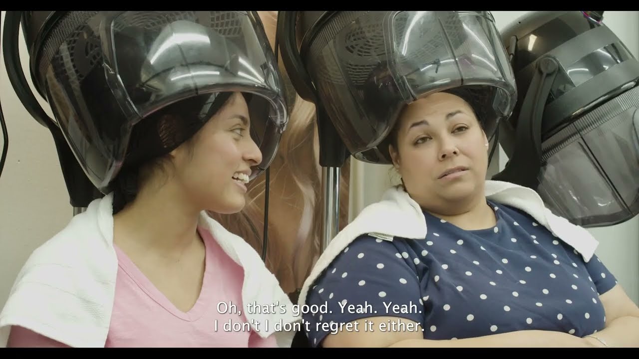 Two Hispanic women get their hair done in a salon and talk with each other.