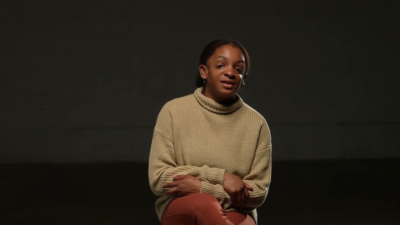 A young Black woman wearing a sweater is speaking. 