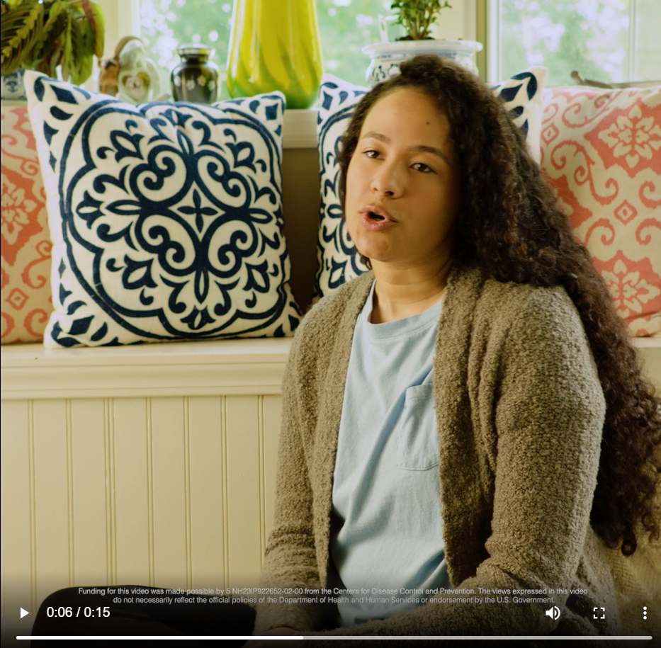 a black woman wearing a light blue t-shirt and a green sweater speaks 