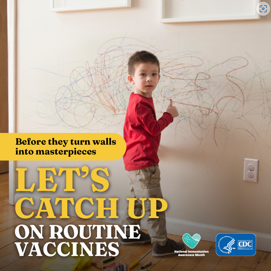 A young boy holding a crayon in his hand stands in front of a wall on which he has scribbled in various colors.