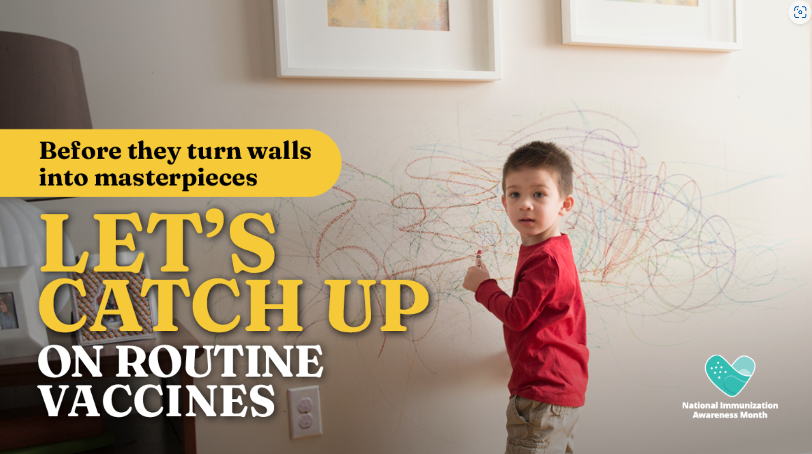 A young boy holding a crayon in his hand stands in front of a wall on which he has scribbled in various colors.