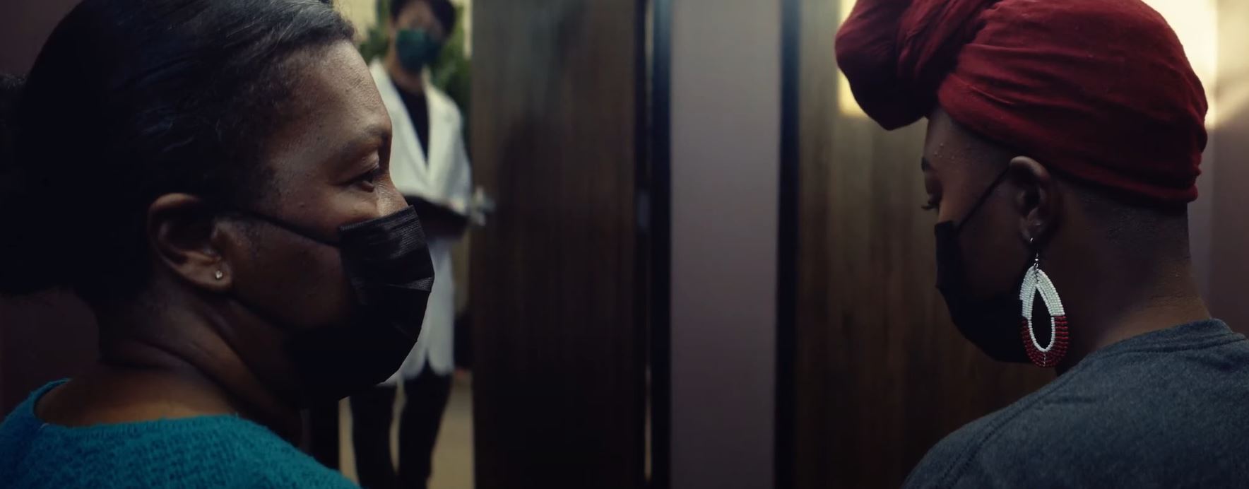 Image of two black women in a doctor's office, wearing masks and looking at each other.