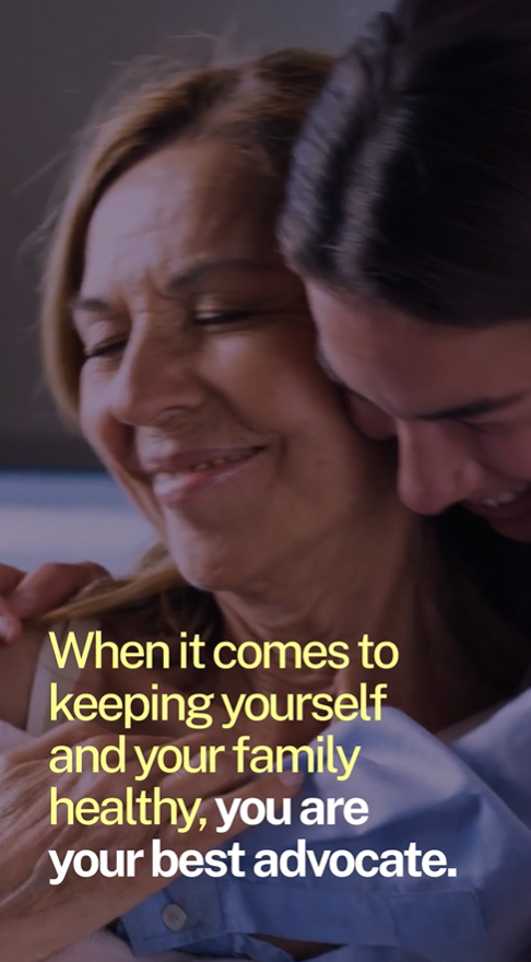 A granddaughter leans over her grandmother's shoulders and hugs her; both have big smiles on their faces.